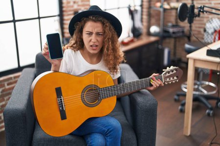 Téléchargez les photos : Jeune femme caucasienne jouant de la guitare classique au studio de musique tenant smartphone en colère et fou hurlant frustré et furieux, criant de colère. rage et concept agressif. - en image libre de droit