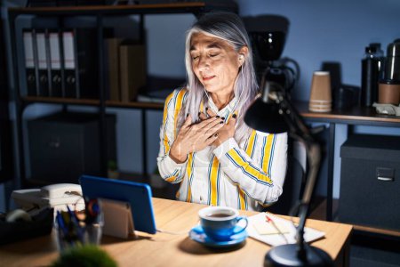 Photo for Middle age woman with grey hair working at the office at night smiling with hands on chest with closed eyes and grateful gesture on face. health concept. - Royalty Free Image