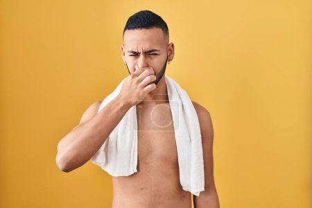 Foto de Joven hispano de pie sin camisa con toalla oliendo algo apestoso y asqueroso, olor intolerable, conteniendo la respiración con los dedos en la nariz. mal olor - Imagen libre de derechos