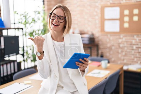 Foto de Young caucasian woman working at the office wearing glasses smiling with happy face looking and pointing to the side with thumb up. - Imagen libre de derechos