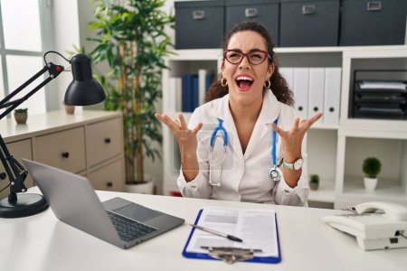 Photo for Young hispanic woman wearing doctor uniform and stethoscope crazy and mad shouting and yelling with aggressive expression and arms raised. frustration concept. - Royalty Free Image