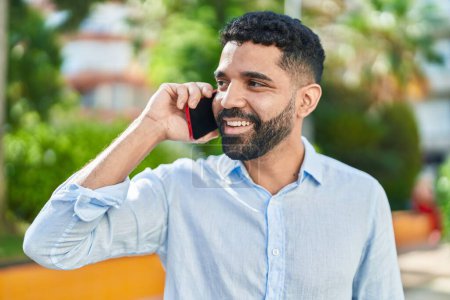 Téléchargez les photos : Jeune homme arabe souriant confiant parler sur le smartphone au parc - en image libre de droit