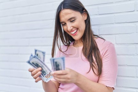 Téléchargez les photos : Young beautiful hispanic woman smiling confident counting dollars over isolated white brick background - en image libre de droit