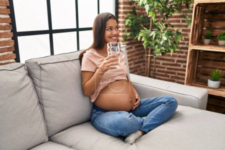 Photo for Young latin woman pregnant drinking water sitting on sofa at home - Royalty Free Image