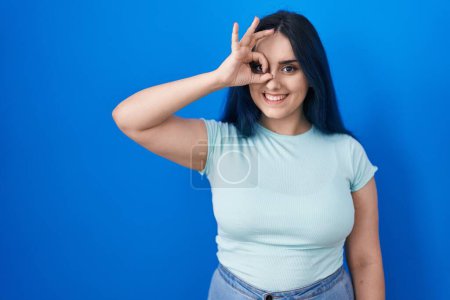 Foto de Young modern girl with blue hair standing over blue background doing ok gesture with hand smiling, eye looking through fingers with happy face. - Imagen libre de derechos