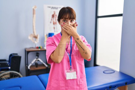Foto de Young brunette woman working at rehabilitation clinic shocked covering mouth with hands for mistake. secret concept. - Imagen libre de derechos