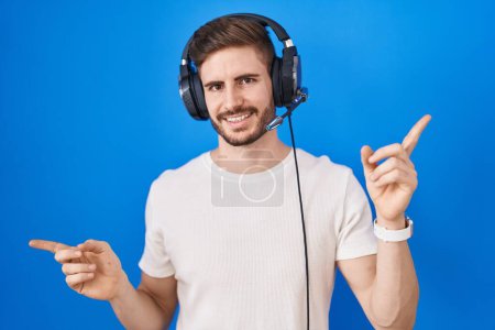Foto de Hombre hispano con barba escuchando música usando auriculares sonriendo confiado señalando con los dedos a diferentes direcciones. espacio de copia para publicidad - Imagen libre de derechos