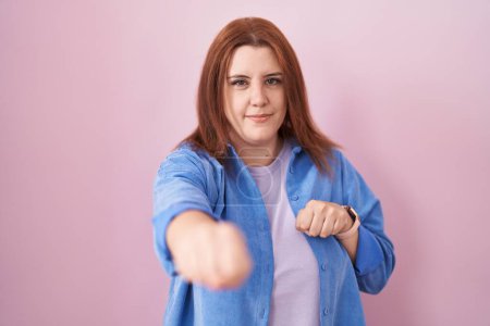 Téléchargez les photos : Young hispanic woman with red hair standing over pink background punching fist to fight, aggressive and angry attack, threat and violence - en image libre de droit