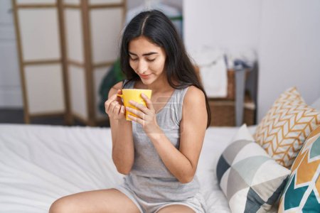 Foto de Young hispanic girl drinking cup of coffee sitting on bed at bedroom - Imagen libre de derechos