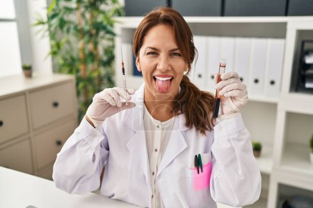 Téléchargez les photos : Hispanic doctor woman holding blood sample and syringe sticking tongue out happy with funny expression. - en image libre de droit
