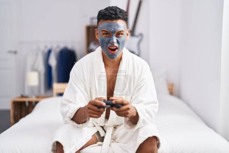 Téléchargez les photos : Young hispanic man wearing beauty face mask playing video games angry and mad screaming frustrated and furious, shouting with anger. rage and aggressive concept. - en image libre de droit