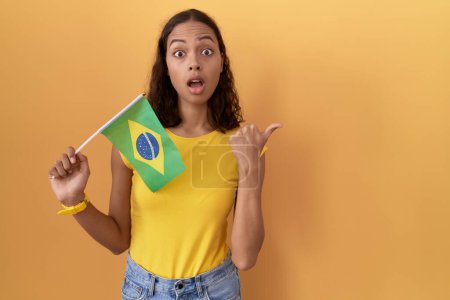 Foto de Young hispanic woman holding brazil flag surprised pointing with hand finger to the side, open mouth amazed expression. - Imagen libre de derechos