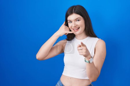 Téléchargez les photos : Jeune femme caucasienne debout sur fond bleu souriant faire parler au téléphone geste et vous pointant du doigt. Appelle-moi. - en image libre de droit