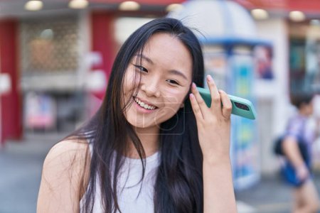 Foto de Joven mujer china sonriendo confiado escuchar mensaje de audio por el teléfono inteligente en la calle - Imagen libre de derechos