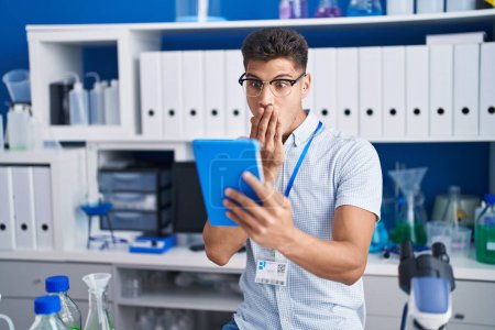 Téléchargez les photos : Young hispanic man working at scientist laboratory doing video call covering mouth with hand, shocked and afraid for mistake. surprised expression - en image libre de droit