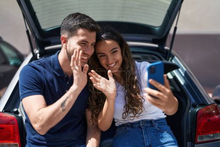 Téléchargez les photos : Jeune couple hispanique assis sur le coffre de la voiture ayant appel vidéo à la rue - en image libre de droit