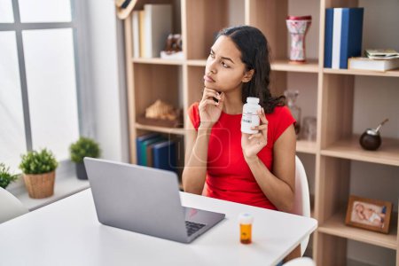 Photo for Young brazilian woman doing video call showing pills to the doctor serious face thinking about question with hand on chin, thoughtful about confusing idea - Royalty Free Image