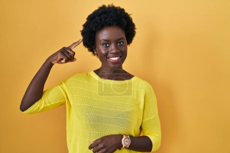 Téléchargez les photos : Jeune femme africaine debout sur un studio jaune souriant pointant vers la tête avec un doigt, bonne idée ou pensée, bonne mémoire - en image libre de droit