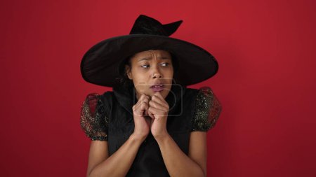 Téléchargez les photos : Femme afro-américaine portant un costume de sorcière faisant geste de peur sur fond rouge isolé - en image libre de droit