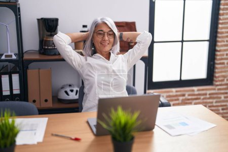 Foto de Trabajadora de negocios de mediana edad de cabello gris usando laptop relajada con las manos en la cabeza en la oficina - Imagen libre de derechos