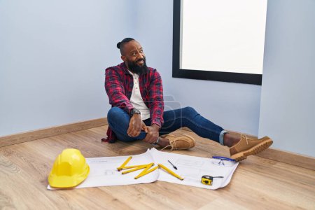 Photo for African american man sitting on the floor at new home looking at blueprints looking away to side with smile on face, natural expression. laughing confident. - Royalty Free Image