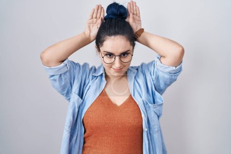 Foto de Young modern girl with blue hair standing over white background doing bunny ears gesture with hands palms looking cynical and skeptical. easter rabbit concept. - Imagen libre de derechos
