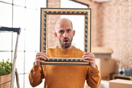 Photo for Young bald man with beard moving to a new home putting face inside vintage frame in shock face, looking skeptical and sarcastic, surprised with open mouth - Royalty Free Image