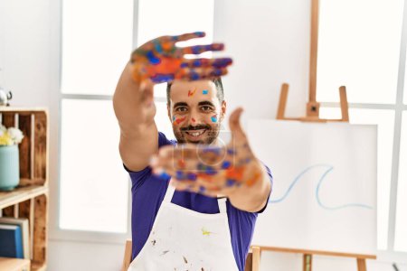 Foto de Joven hispano sonriendo confiado mostrando las manos pintadas de palma en el estudio de arte - Imagen libre de derechos