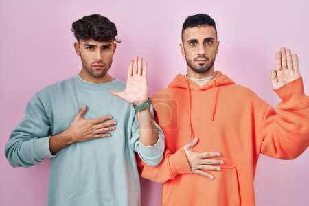 Foto de Young hispanic gay couple standing over pink background swearing with hand on chest and open palm, making a loyalty promise oath - Imagen libre de derechos
