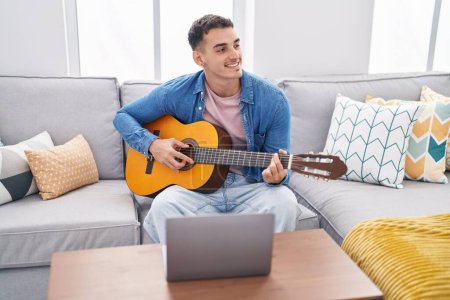 Photo for Young hispanic man having online classical guitar lesson sitting on sofa at home - Royalty Free Image