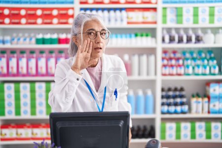 Foto de Middle age woman with tattoos working at pharmacy drugstore hand on mouth telling secret rumor, whispering malicious talk conversation - Imagen libre de derechos