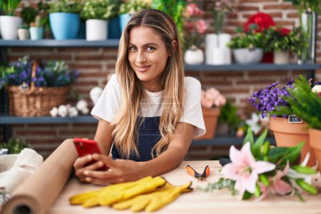 Téléchargez les photos : Jeune femme blonde fleuriste souriant confiant en utilisant un smartphone chez le fleuriste - en image libre de droit
