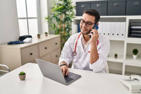 Photo for Young hispanic man wearing doctor uniform using laptop talking on the smartphone at clinic - Royalty Free Image