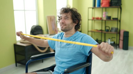 Foto de Young hispanic man sitting on wheelchair stretching arms with elastic band at rehab clinic - Imagen libre de derechos
