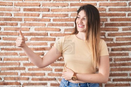 Téléchargez les photos : Jeune femme brune debout au-dessus du mur de briques regardant fier, souriant faire les pouces vers le haut geste sur le côté - en image libre de droit