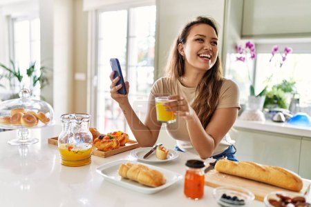 Photo for Young beautiful hispanic woman having breakfast using smartphone at the kitchen - Royalty Free Image