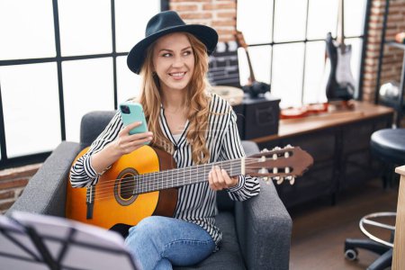 Foto de Joven mujer rubia músico usando smartphone sosteniendo la guitarra clásica en el estudio de música - Imagen libre de derechos