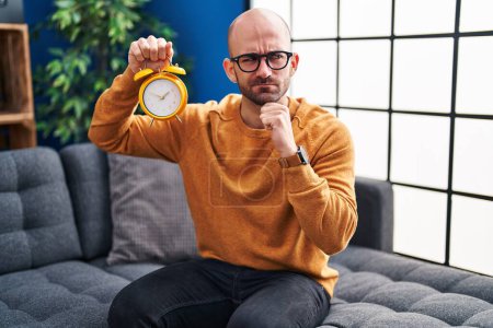Photo for Young bald man with beard holding alarm clock serious face thinking about question with hand on chin, thoughtful about confusing idea - Royalty Free Image