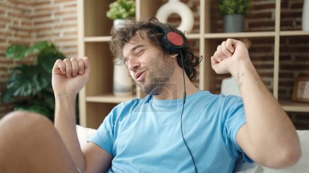 Photo for Young hispanic man sitting on bed listening to music and dancing at bedroom - Royalty Free Image