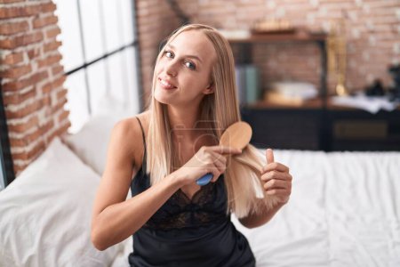 Foto de Young blonde woman combing hair sitting on bed at bedroom - Imagen libre de derechos