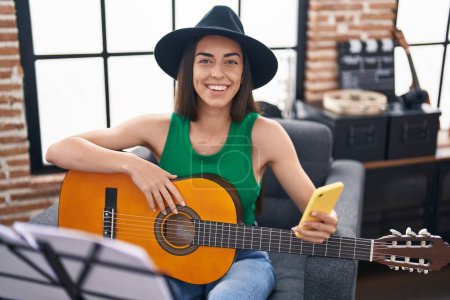 Photo for Young hispanic woman musician using smartphone playing classical guitar at music studio - Royalty Free Image