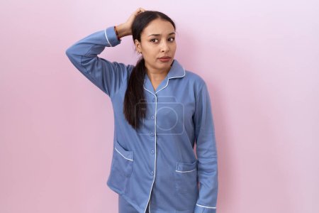Foto de Young arab woman wearing blue pajama confuse and wondering about question. uncertain with doubt, thinking with hand on head. pensive concept. - Imagen libre de derechos