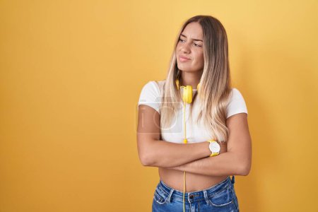 Téléchargez les photos : Young blonde woman standing over yellow background wearing headphones looking to the side with arms crossed convinced and confident - en image libre de droit