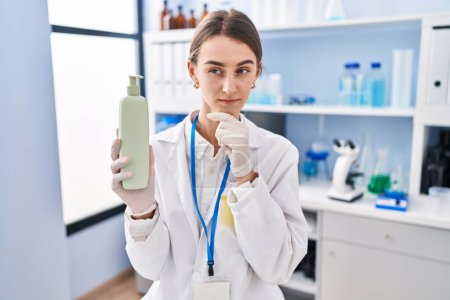 Photo for Young caucasian woman working at scientist laboratory holding body lotion serious face thinking about question with hand on chin, thoughtful about confusing idea - Royalty Free Image