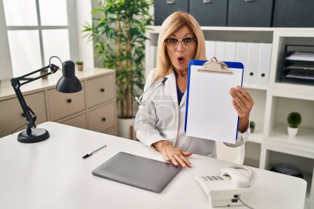 Téléchargez les photos : Middle age blonde doctor woman holding clipboard at the clinic scared and amazed with open mouth for surprise, disbelief face - en image libre de droit
