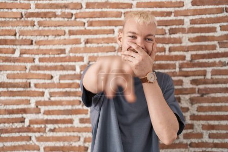 Photo for Young caucasian man standing over bricks wall laughing at you, pointing finger to the camera with hand over mouth, shame expression - Royalty Free Image