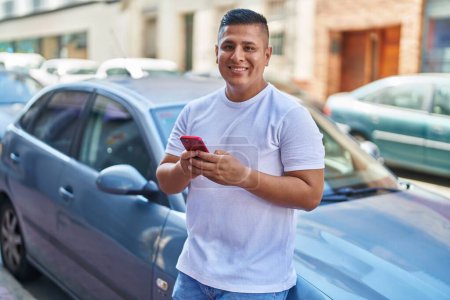 Téléchargez les photos : Jeune homme latin en utilisant smartphone appuyé sur la voiture à la rue - en image libre de droit