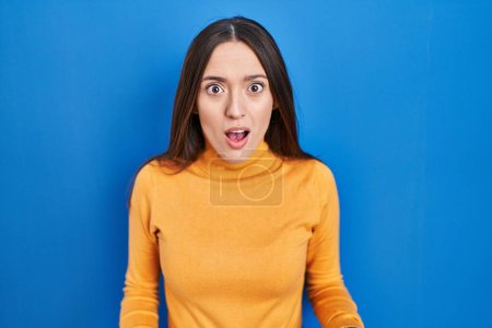 Photo for Young brunette woman standing over blue background afraid and shocked with surprise and amazed expression, fear and excited face. - Royalty Free Image