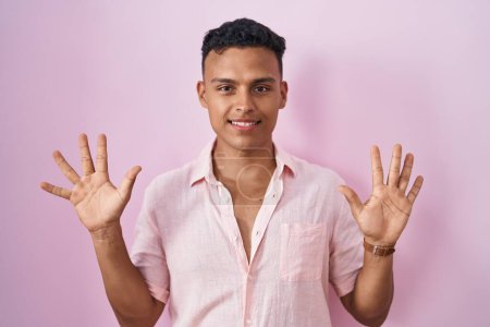 Foto de Joven hombre hispano de pie sobre fondo rosa mostrando y señalando hacia arriba con los dedos número diez mientras sonríe confiado y feliz. - Imagen libre de derechos