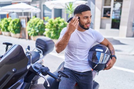 Photo for Young latin man listening voice message by smartphone sitting on motorbike at street - Royalty Free Image